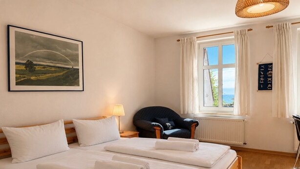 Et hotelværelse med seng, stol, et vindue med gardiner, en lampe og et billede i ramme på væggen.