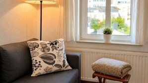 Et værelse med en sofa, en lampe, et vindue med udsigt til bygninger og en lille plante på vindueskarmen.