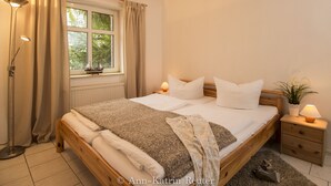 Ein ordentlich gemachtes Bett mit Holzrahmen, ein Nachttisch mit Lampe und ein Fenster mit Vorhängen.