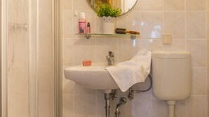 Ein Badezimmer mit Waschbecken, Toilette und Spiegel. Auf dem Waschbecken steht eine Pflanze, ein Handtuch hängt an der Wand und ein Paar Hausschuhe liegt auf dem Boden.