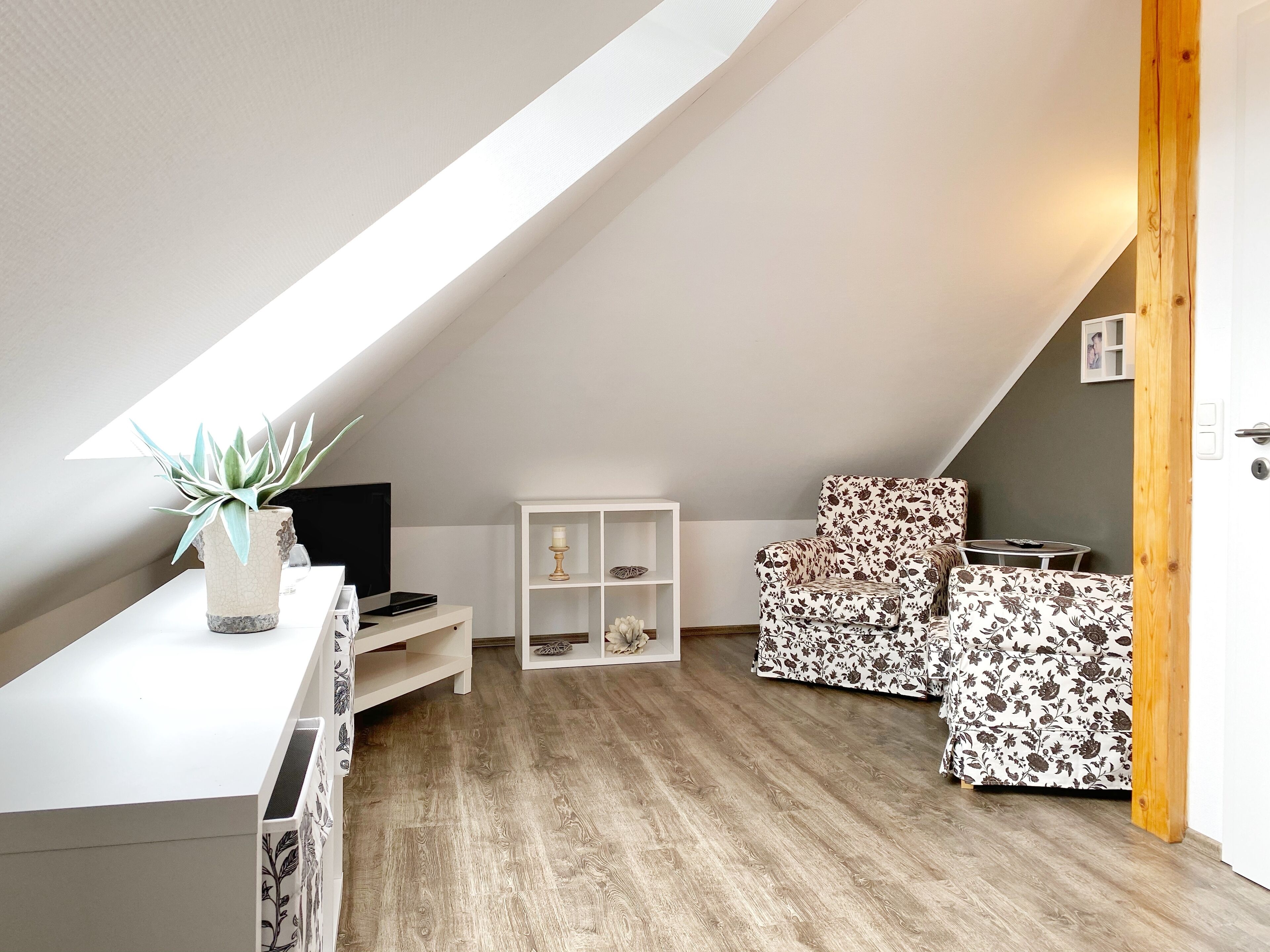 A cozy attic bedroom with a sloped ceiling, wooden flooring, a white desk with a plant, a TV, and a floral-patterned sofa.
