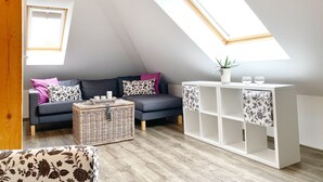 A loft-style room with a sloped ceiling, a dark grey sofa with floral cushions, a wicker basket, a white shelving unit with floral fabric, and a small potted plant. - Ferienhaus "sabrina" (Sassnitz)