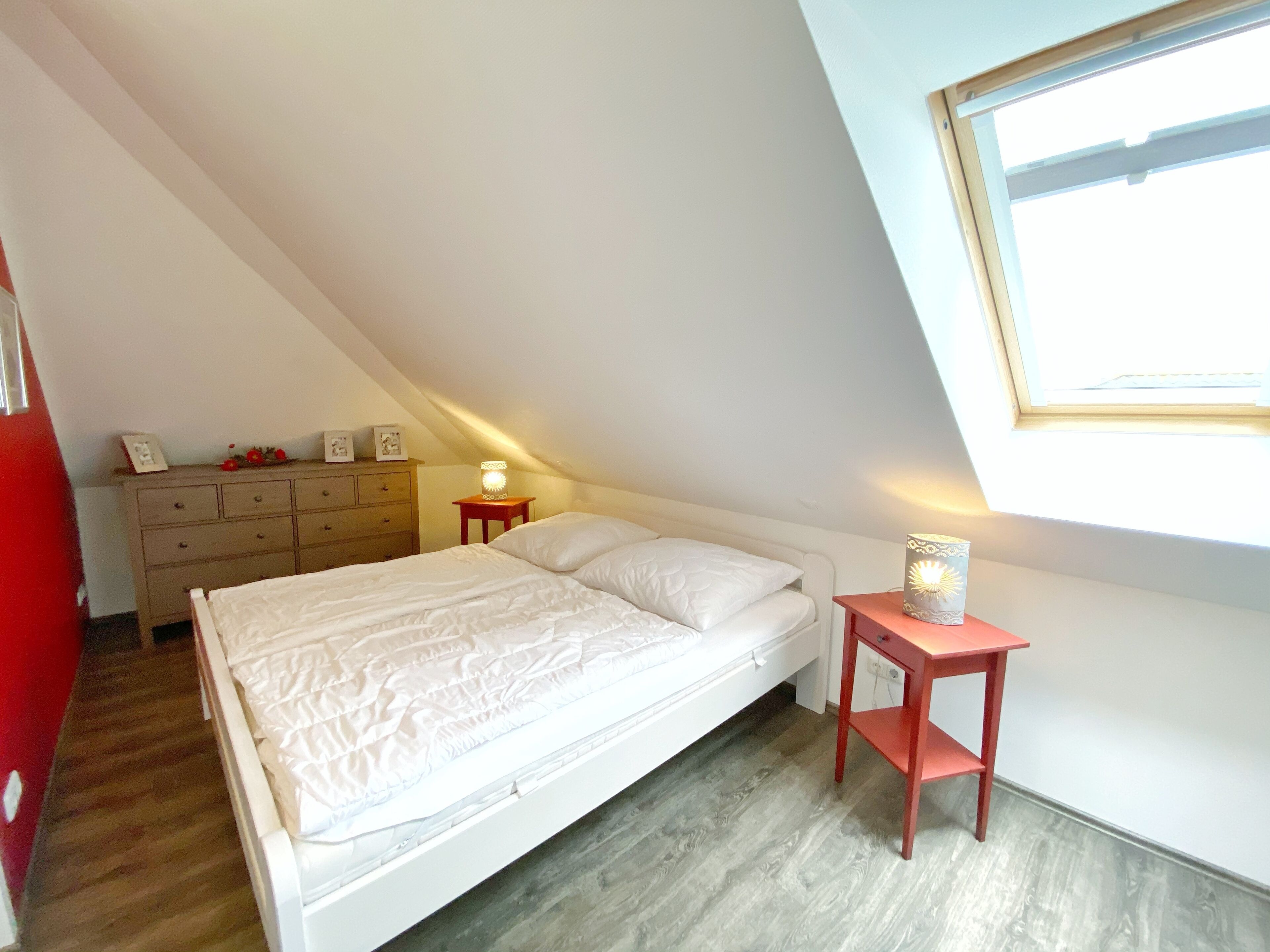 Een slaapkamer met een schuine kap, een bed, een dressoir, een nachtkastje met een lamp en een raam.