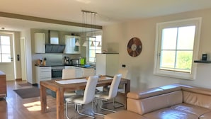A modern living room with a wooden dining table, white chairs, a beige leather sofa, and a kitchen in the background. - Ferienhaus "sarcander" (Glowe)