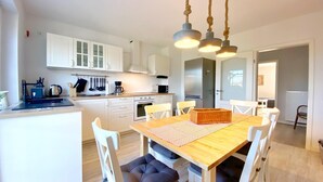 A modern kitchen with white cabinets, a central island, and a dining area with a wooden table and white chairs. - Reethaus Vilmnitz - Ferienwohnung "muglitz" (Vilmnitz)