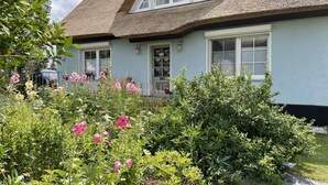 A house with a thatched roof, a front porch, and a garden with pink flowers. - Reethaus Vilmnitz - Ferienwohnung "muglitz" (Vilmnitz)