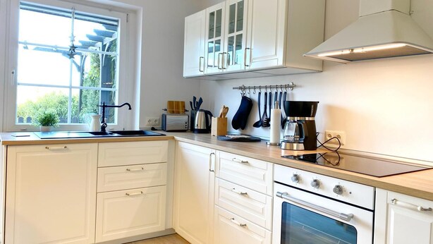 Una cocina moderna con armarios blancos, horno integrado, cafetera y ventana con vista a follaje.
