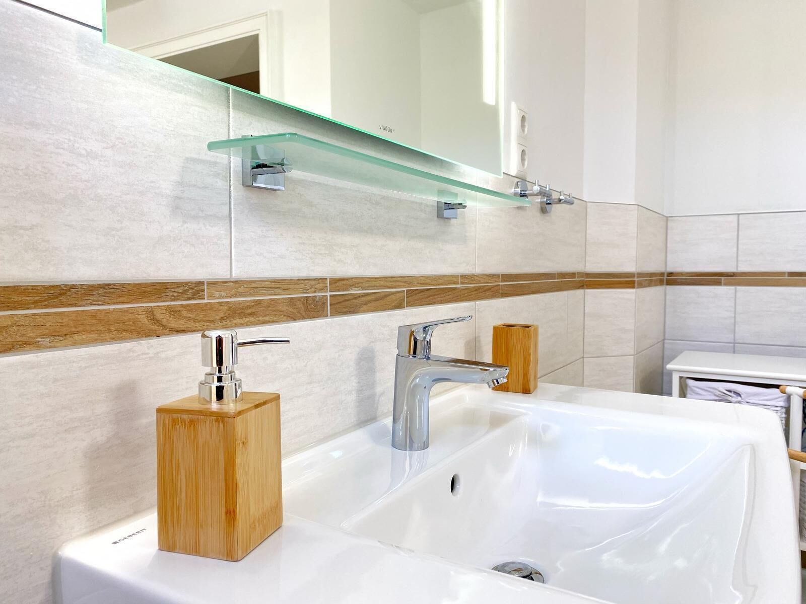 Une salle de bain moderne avec un lavabo blanc, deux robinets, un distributeur de savon en bois et une étagère en verre au-dessus du lavabo.