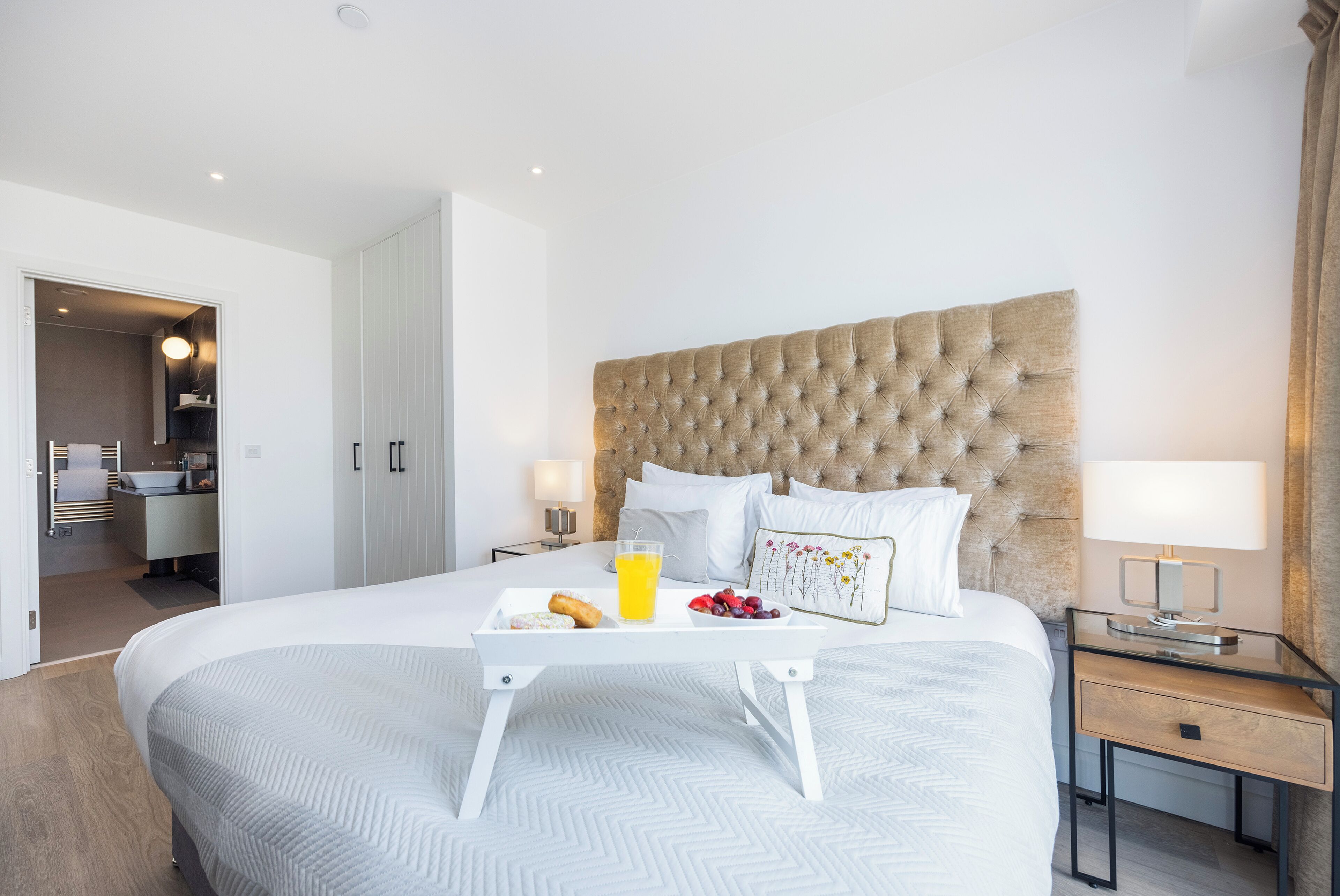 A modern hotel room with a large bed, a bedside table with a lamp, and a tray with breakfast items.