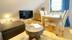 Una sala de estar acogedora con un televisor de pantalla plana, una mesa de comedor de madera con sillas, un sofá y una pequeña mesa redonda con una vela.