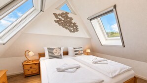 Un dormitorio con techo inclinado, ventanas de tragaluces, una cama con ropa blanca, dos lámparas de noche y una mesita de noche de madera.