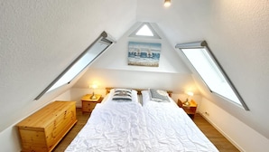 Un dormitorio con techo inclinado, tragaluces, una cama con ropa de cama blanca, dos mesitas de noche con lámparas y un aparador de madera.