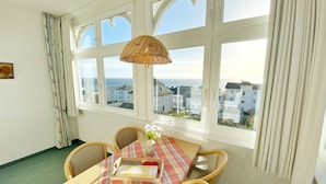 Un coin repas avec une table dressée pour deux personnes, une lampe en osier et une vue sur la mer grâce à de grandes fenêtres.