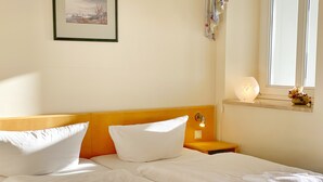 Un letto rifatto con lenzuola e cuscini bianchi, un comodino con una lampada e una finestra con vista sull'esterno.