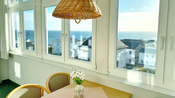 Una habitación con ventanas amplias que dan a una ciudad costera, una lámpara de mimbre, una mesa con un florero y sillas de mimbre.