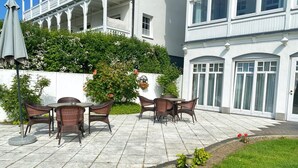 Un patio con mobiliario de mimbre, un edificio blanco con balcones y un jardín lleno de flores.