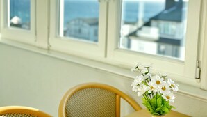 Una habitación con ventana que da a la costa, una mesa con un florero y una silla.