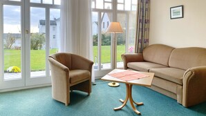 A room with a sofa, armchair, and a table with a placemat. There are large windows with curtains, a lamp, and a framed picture on the wall. - Ferienappartement 10 in der Villa Anna in Sassnitz (Sassnitz)