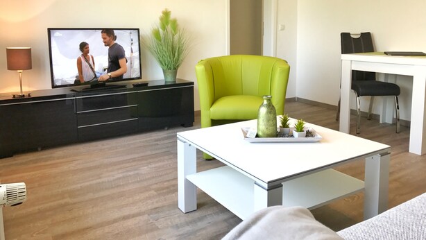 Una sala de estar moderna con un televisor de pantalla plana, un sillón verde, una mesa de centro blanca y una zona de comedor con una mesa blanca y sillas negras.