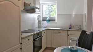 Una cocina compacta con armarios blancos, horno integrado, cafetera y ventana con persianas.