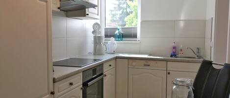 Una cocina compacta con armarios blancos, horno integrado, cafetera y ventana con persianas.