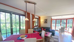 A spacious living area with a dining table, chairs, a sofa, and large windows offering a view of greenery.