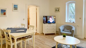 Ein Wohnzimmer mit Esstisch, Stühlen, Fernseher, Sofa, Fenster und einer dekorativen Deckenleuchte.