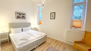 Ein Schlafzimmer mit einem Bett, einem Nachttisch, einem Sessel, einem Heizkörper und zwei Fenstern.