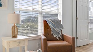 Una habitación con ventana, una silla con manta, una lámpara y una puerta.