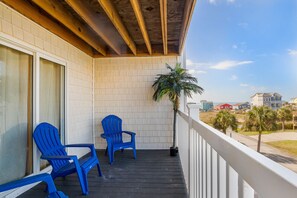 A balcony with two blue chairs, a potted palm tree, and a view of the beach and buildings.