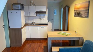 A compact kitchen with white cabinets, a refrigerator, and a coffee maker. There's a dining table with chairs and a painting on the wall.