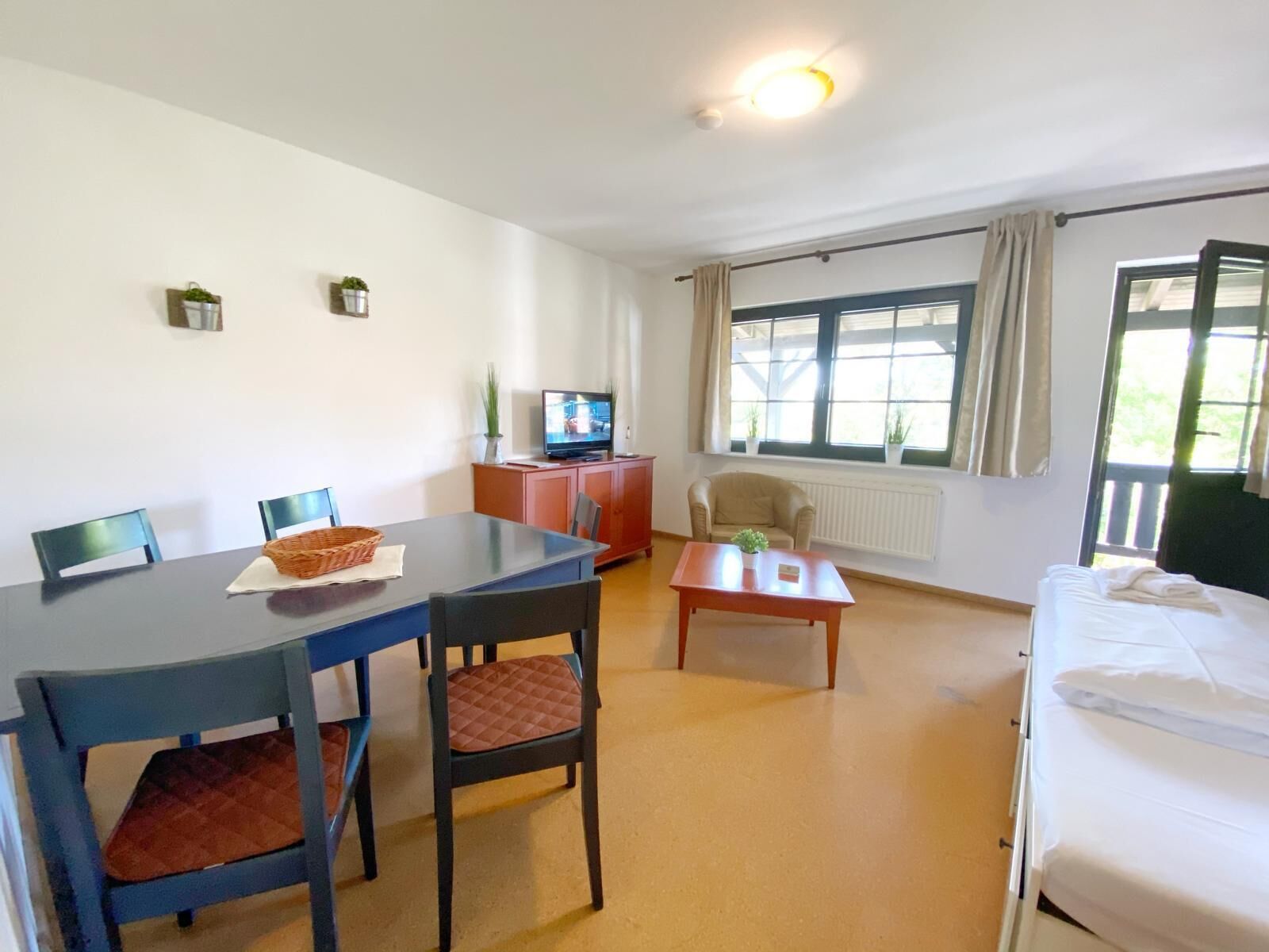 A room with a bed, a desk with a computer, a chair, a table, a TV, and a window with curtains.