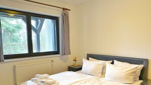 A neatly made bed with white linens and towels, a bedside table with a lamp, a window with checkered curtains, and a radiator. - Landpension Dubnitz - Holiday apartment 2 (Sassnitz)
