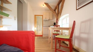 Un salon chaleureux avec un canapé rouge, une table à manger en bois et deux chaises, et une cuisine en arrière-plan.