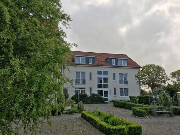 Un bâtiment à deux étages, doté d’un toit recouvert de tuiles rouges, de nombreuses fenêtres et d’un jardin bien entretenu avec des haies et un banc.