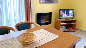 Ruokailutila, jossa on puinen pöytä, tuoleja, punottu kori, takka, televisio ja kirjahylly.