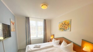 Hotellihuone, jossa on suuri sänky, yöpöytälamppu, seinälle asennettu televisio ja ikkuna, jossa on verhot.