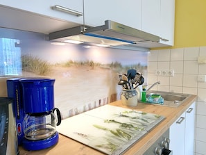 Una cocina con cafetera, tabla de cortar con vegetales, lavamanos y campana extractora fijada a la pared.
