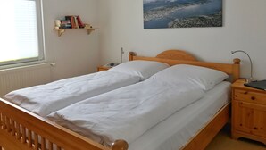A bedroom with a wooden bed, white bedding, a nightstand with a lamp, a shelf with books, and a painting of a landscape. - Hochzeitsberg - Double room / Apartment "Tromsö" in Breege (Breege)