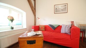 Ein gemütliches Wohnzimmer mit einem roten Sofa, einer Holzkommode, einem Fenster mit Blumen in einer Vase und einem gerahmten Bild an der Wand.