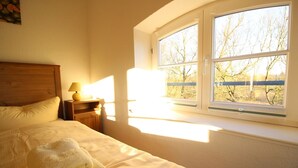 Ein Schlafzimmer mit Bett, Nachttisch, Lampe und Fenster mit Blick auf Bäume.