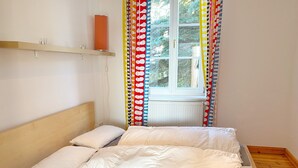 A single bed with a wooden headboard, white bedding, and a window with colorful curtains.