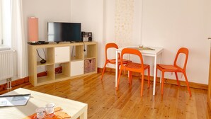 A room with a wooden floor, a white table with orange chairs, a TV on a wooden shelf, and a radiator.