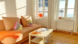 A cozy living room with a wooden floor, a beige sofa with patterned cushions, a white coffee table, and a window with a view of the sea.