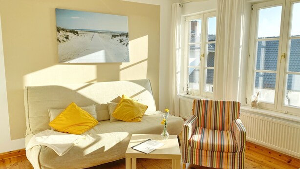 A living room with a beige sofa, yellow cushions, a striped armchair, a small table with a vase of flowers, and a painting of a beach scene on the wall. - Seaside Appartements, "yellow" (Sassnitz)