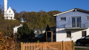 Une maison moderne à deux étages, dotée d’un balcon, entourée d’une clôture en bois et d’arbres.