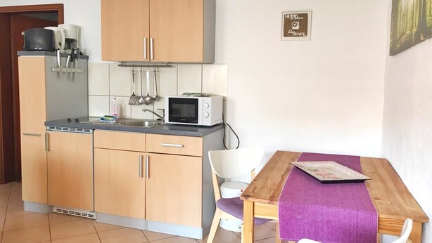 Cocina pequeña con armarios de madera, microondas y pileta. Hay una mesa de comedor con camino de mesa morado y sillas blancas.