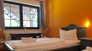 A bedroom with two beds, a window with curtains, a bedside table with a lamp, and a view of buildings outside.