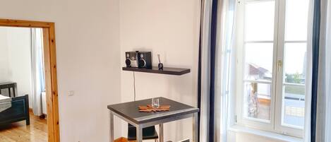 Ein kleiner, moderner Raum mit Holzboden, einem Tisch mit zwei Stühlen, einem Regal mit Radio und Glas sowie einem Fenster mit Vorhängen.