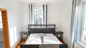 A bedroom with a wooden floor, a bed with white bedding, two bedside tables, a window with curtains, and a radiator. - Seaside Apartments, "Brown" (Sassnitz)